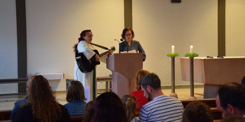 2 Personen mit einer Gitarre stehen vor einem Publikum an einem Pult